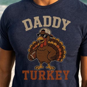 May include: A navy blue short-sleeved t-shirt featuring a graphic of a turkey wearing a baseball cap and sunglasses while holding a football. The text DADDY is printed in light beige above the turkey, and the word TURKEY is printed in orange below.
