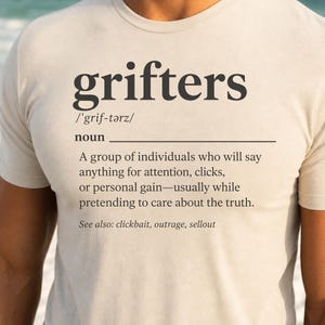 May include: A beige crew neck t-shirt featuring a dictionary-style definition of the word grifters. The text includes the phonetic spelling, the noun classification, and a descriptive definition about individuals seeking attention or personal gain.