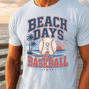May include: A light blue t-shirt featuring a graphic print with the text BEACH DAYS & BASEBALL. The design includes a baseball, palm trees, a sunset over the ocean, and the text USA EST. 1987. The lettering is in dark blue and red.