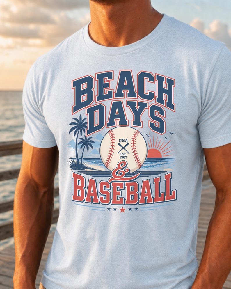 May include: A light blue t-shirt featuring a graphic print with the text BEACH DAYS & BASEBALL. The design includes a baseball, palm trees, a sunset over the ocean, and the text USA EST. 1987. The lettering is in dark blue and red.