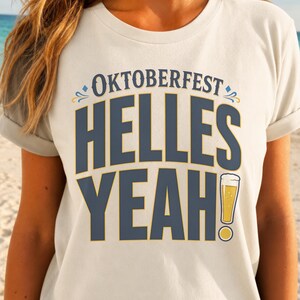 May include: A cream-colored short-sleeve t-shirt featuring the text "OKTOBERFEST" in blue above the large blue text "HELLES YEAH!" with a graphic of a glass of golden beer replacing the exclamation point, accented by small blue and yellow flourishes.