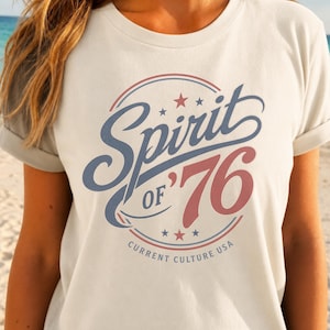 May include: A cream-colored short-sleeve t-shirt featuring a circular graphic with the blue script text Spirit, the red text OF '76, and the small text CURRENT CULTURE USA. The design includes red and blue stars and circular border lines.