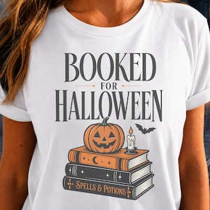 May include: A white t-shirt featuring the text BOOKED FOR HALLOWEEN in dark gray. Below the text is a graphic of a stack of three books, a carved orange jack-o'-lantern, a lit candle, and a small black bat. The bottom book is labeled SPELLS & POTIONS.