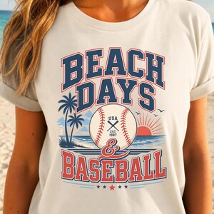 May include: A cream-colored t-shirt featuring a graphic with the text BEACH DAYS & BASEBALL. The design includes a baseball, palm trees, a setting sun over the ocean, and the text USA EST. 1987. The color palette consists of navy blue and red.