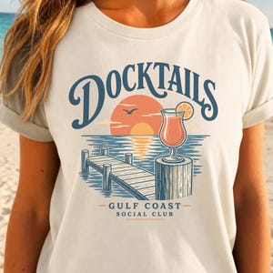 May include: A cream-coloured t-shirt featuring a graphic of a wooden dock, a cocktail glass with an orange slice, and a sunset over water. The text reads DOCKTAILS, GULF COAST, and SOCIAL CLUB in blue and orange lettering on the front of the shirt.