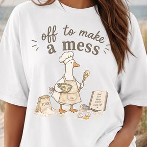 May include: A white t-shirt featuring a graphic of a goose wearing a chef hat and apron, holding a mixing bowl and spoon. The text reads "off to make a mess" above the goose, with a bag of flour, a whisk, and an open cookbook labeled "Good Food Messy Kitchen Happy Life" below.