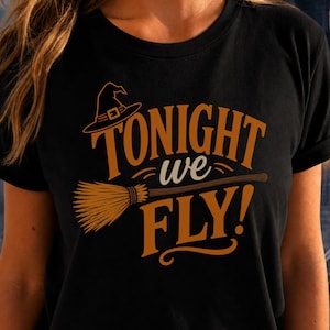 May include: A black short-sleeved t-shirt featuring a graphic print in orange and cream. The design includes a witch hat, a broomstick, and the text "TONIGHT we FLY!" in a bold, stylized font centered on the front of the garment.