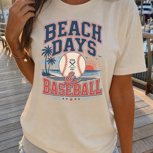 May include: A cream-colored short-sleeve t-shirt featuring a graphic print with the text BEACH DAYS & BASEBALL. The design includes a baseball, palm trees, a sunset over the ocean, and the text USA EST. 1987 centered on the baseball.