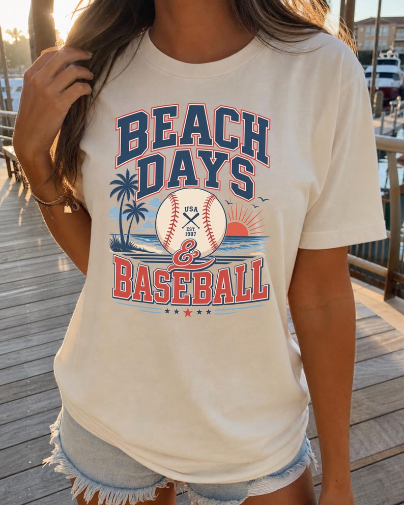 May include: A cream-colored short-sleeve t-shirt featuring a graphic print with the text BEACH DAYS & BASEBALL. The design includes a baseball, palm trees, a sunset over the ocean, and the text USA EST. 1987 centered on the baseball.