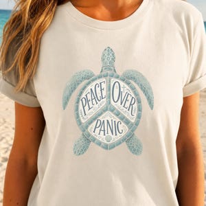 May include: A cream-colored short-sleeve t-shirt featuring a graphic of a sea turtle. The turtle's shell is divided into three sections containing the text "PEACE OVER PANIC" in a dark blue font, accented with small decorative floral and shell motifs.