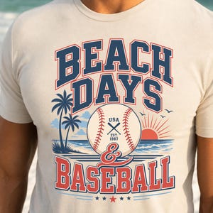 May include: A cream-colored short-sleeve t-shirt featuring a graphic print with the text BEACH DAYS & BASEBALL. The design includes a baseball, palm trees, a sunset over the ocean, and the text USA EST. 1987, rendered in navy blue and red.