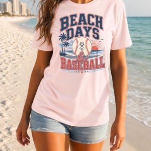 May include: A light pink short-sleeve t-shirt featuring a graphic print with the text BEACH DAYS & BASEBALL. The design includes a baseball, palm trees, a rising sun, and ocean waves in blue, red, and white colors, with USA EST 1987 on the ball.