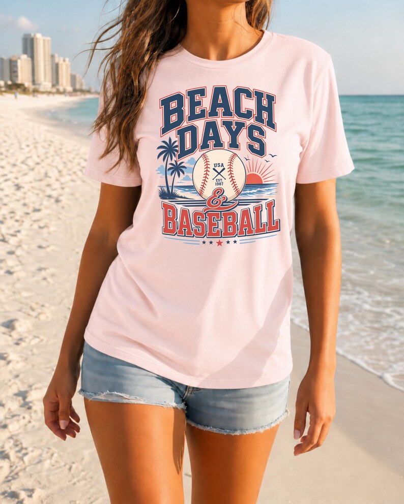 May include: A light pink short-sleeve t-shirt featuring a graphic print with the text BEACH DAYS & BASEBALL. The design includes a baseball, palm trees, a rising sun, and ocean waves in blue, red, and white colors, with USA EST 1987 on the ball.