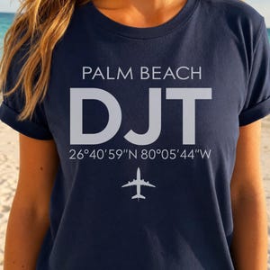 May include: A navy blue short-sleeved t-shirt featuring the white text PALM BEACH, the letters DJT, the coordinates 26°40'59"N 80°05'44"W, and a small white airplane icon centered on the chest area of the garment.