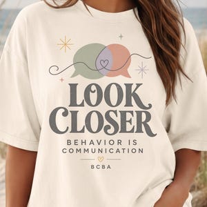 Könnte beinhalten: Ein cremefarbenes T-Shirt mit kurzen Ärmeln und einer Grafik aus zwei überlappenden Sprechblasen in Salbeigrün und gedecktem Rosa. Der Text lautet LOOK CLOSER, BEHAVIOR IS COMMUNICATION und BCBA, verziert mit kleinen Sternen und einem Herz.
