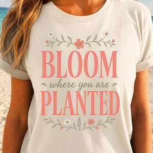 May include: A cream-colored short-sleeve t-shirt featuring a graphic print with the text "BLOOM where you are PLANTED" in coral pink lettering. The design is framed by delicate green stems, leaves, and small white and coral pink flowers.
