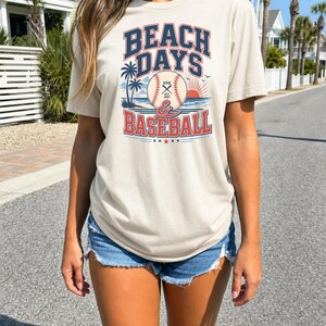 May include: A beige short-sleeve t-shirt featuring a graphic print with the text BEACH DAYS & BASEBALL. The design includes a baseball, palm trees, a sun, and birds. The text BEACH DAYS is in dark blue, and BASEBALL is in red.