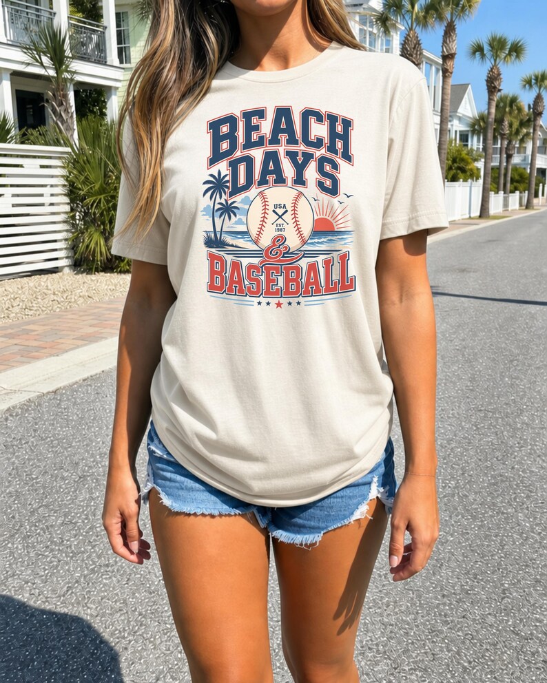 May include: A beige short-sleeve t-shirt featuring a graphic print with the text BEACH DAYS & BASEBALL. The design includes a baseball, palm trees, a sun, and birds. The text BEACH DAYS is in dark blue, and BASEBALL is in red.