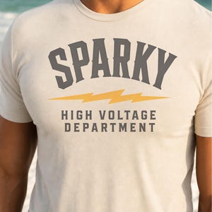 May include: A beige short-sleeved t-shirt featuring the dark gray text SPARKY in an arched font above a yellow lightning bolt graphic. Below the lightning bolt, the text HIGH VOLTAGE DEPARTMENT is printed in dark gray capital letters.