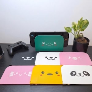 May include: A collection of colorful, rectangular console dock faceplates featuring various animal faces, including a teal turtle, black cat, white dog, pink pig, pink axolotl, yellow chick, and panda. One teal faceplate is attached to a game console.