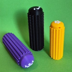 Op de afbeelding: Drie cilindrische massagetools in paars, zwart en geel, elk met een textuuroppervlak van kleine, gelijkmatig verdeelde uitsteeksels. De paarse en gele tools hebben witte decoratieve accenten aan de uiteinden. De achtergrond is effen groen.
