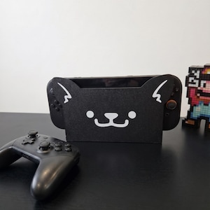 Peut inclure: Une station d'accueil pour console de jeux vidéo noire ornée d'un motif de visage de chat blanc avec des oreilles pointues, deux yeux circulaires et une petite bouche. Une manette sans fil noire est au premier plan et un personnage pixelisé.