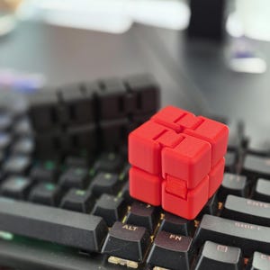 Può includere: Un cubo fidget rosso è appoggiato su una tastiera nera, con un secondo cubo fidget nero sullo sfondo. Il cubo rosso è a fuoco, mostrando il suo design modulare. I tasti della tastiera sono neri con lettere bianche.