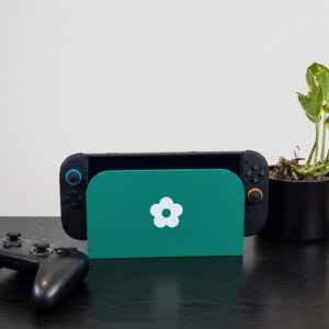 May include: A handheld gaming console docked in a dark green stand featuring a white flower icon. The console has black controllers with one blue and one orange joystick. A separate black wireless game controller sits to the left of the dock.