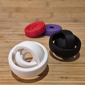 Puede incluir: Cuatro objetos geométricos impresos en 3D de colores. Una forma blanca, similar a una cinta de Möbius, se encuentra en primer plano, con un objeto negro en forma de anillo a la derecha. Dos objetos circulares en forma de disco, rojos y morados, están en el fondo.