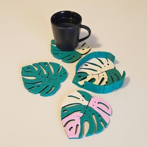 Peut inclure: Une tasse noire repose sur un dessous de verre en forme de feuille de monstera vert et blanc. D'autres dessous de verre verts, blancs et roses sont présentés. Les dessous de verre sont conçus pour ressembler à des feuilles de plantes tropicales.