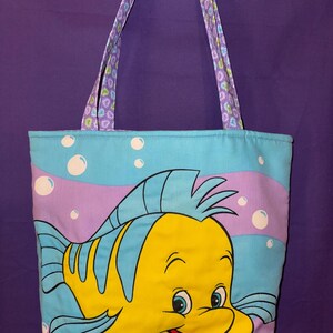 May include: A rectangular fabric tote bag featuring a large, colorful graphic of a yellow and blue fish character. The background is light blue with purple wavy stripes and white bubbles. The bag has two shoulder straps with a purple floral pattern.