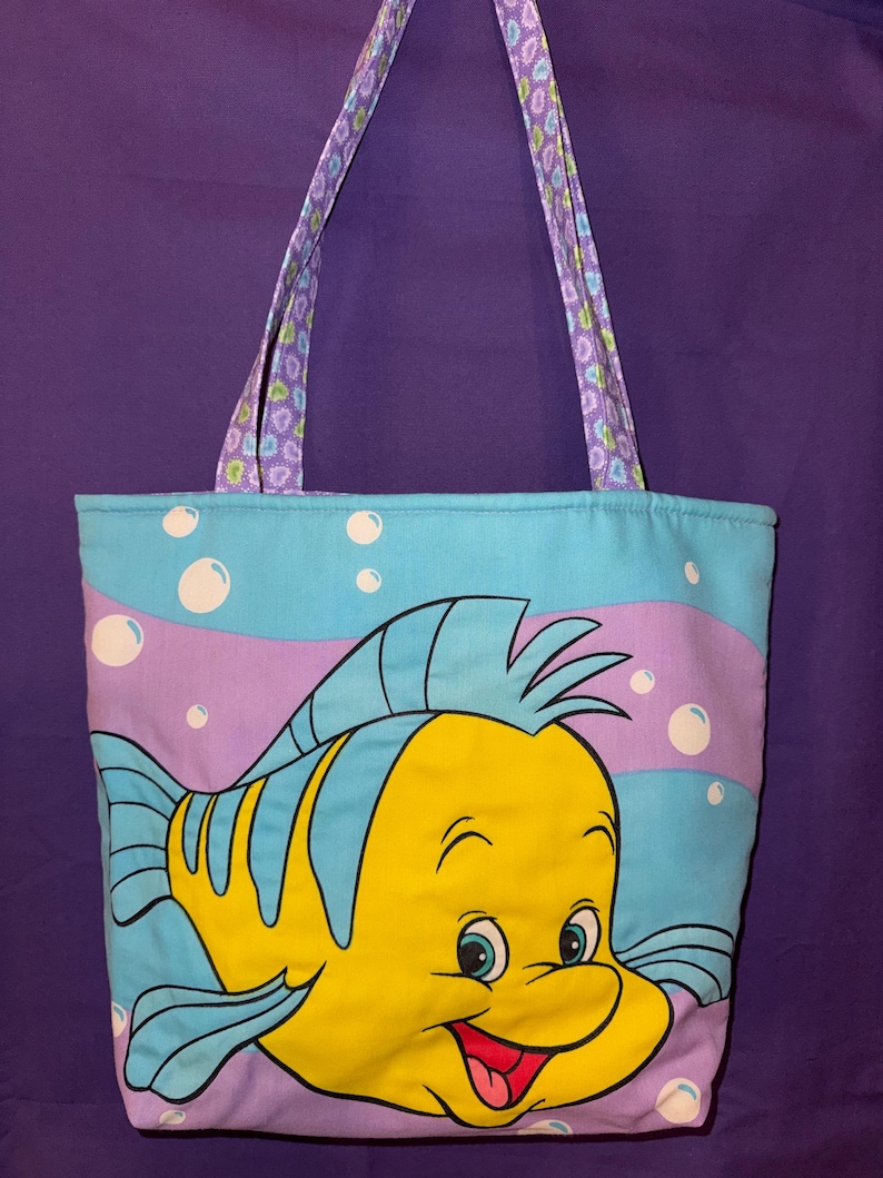 May include: A rectangular fabric tote bag featuring a large, colorful graphic of a yellow and blue fish character. The background is light blue with purple wavy stripes and white bubbles. The bag has two shoulder straps with a purple floral pattern.