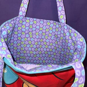 May include: A fabric tote bag featuring a cartoon character with bright red hair on the exterior. The interior lining and the two shoulder straps are decorated with a repeating pattern of small blue and green hearts on a purple background.
