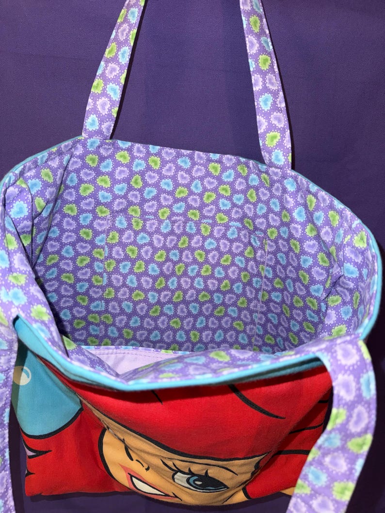 May include: A fabric tote bag featuring a cartoon character with bright red hair on the exterior. The interior lining and the two shoulder straps are decorated with a repeating pattern of small blue and green hearts on a purple background.