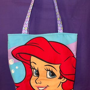 May include: A fabric tote bag featuring a large graphic of a red-haired character with blue eyes on a light blue background with white bubbles. The bag has two shoulder straps made of a purple fabric with a small, multi-colored heart pattern.
