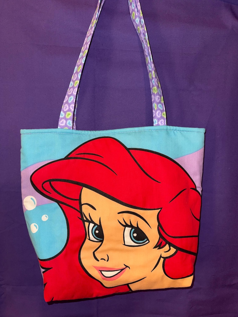 May include: A fabric tote bag featuring a large graphic of a red-haired character with blue eyes on a light blue background with white bubbles. The bag has two shoulder straps made of a purple fabric with a small, multi-colored heart pattern.