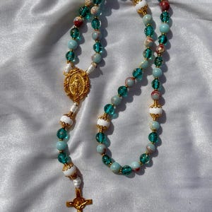 May include: A rosary with turquoise, white, and red beads, accented with gold-colored metal. The rosary features a gold-colored crucifix and a gold-colored medallion with a religious figure.