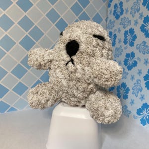 Könnte beinhalten: Ein handgefertigtes Otter-Plüschtier in Grau- und Weißtönen mit schwarzen Augen und Nase. Der Otter sitzt auf einem weißen Kunststoffbehälter, mit einem blau-weiß gemusterten Hintergrund.