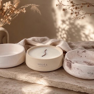 May include: Three round, low-profile vessels made of a matte, stone-like material. One is plain off-white, one is a beige candle with a single dark wick and the text SOLÉA, and one features a marbled brown and white pattern with the text SOLÉA.