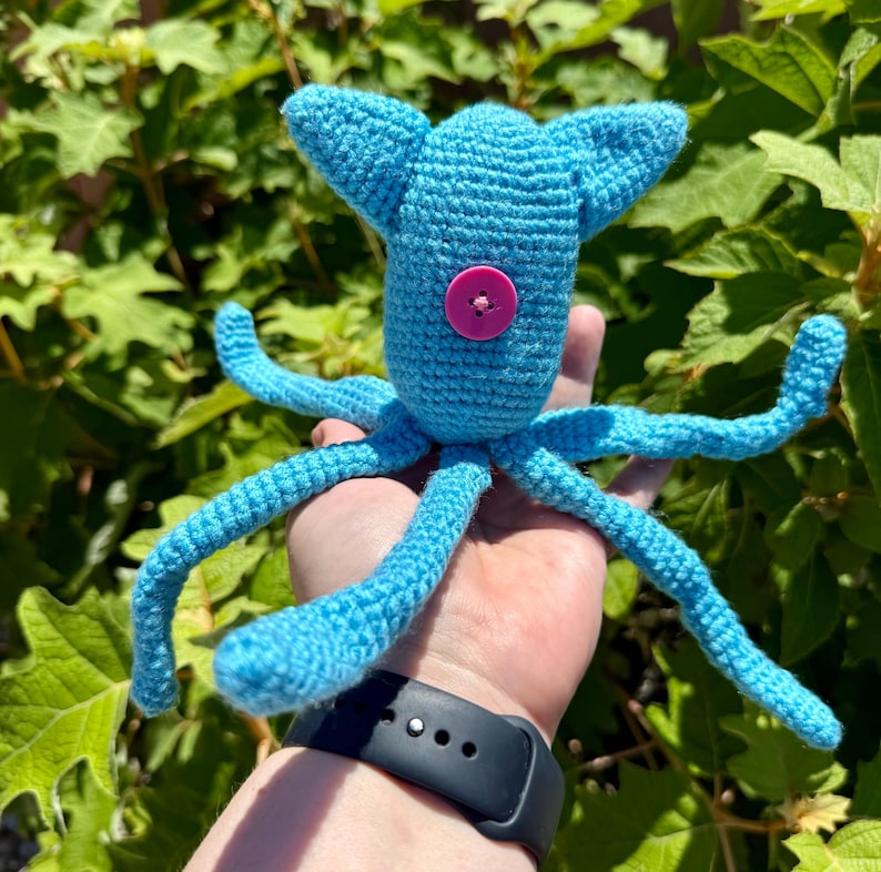 May include: A bright blue crocheted squid toy featuring a single pink button eye centered on its head. The creature has a rounded head with two pointed top fins and eight long, slender, flexible tentacles extending outward from the base.