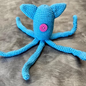 May include: A bright blue crocheted octopus toy featuring a central pink four-hole button on its main body. The creature has two pointed ears at the top and eight long, slender, flexible tentacles extending outward from the base of its head.