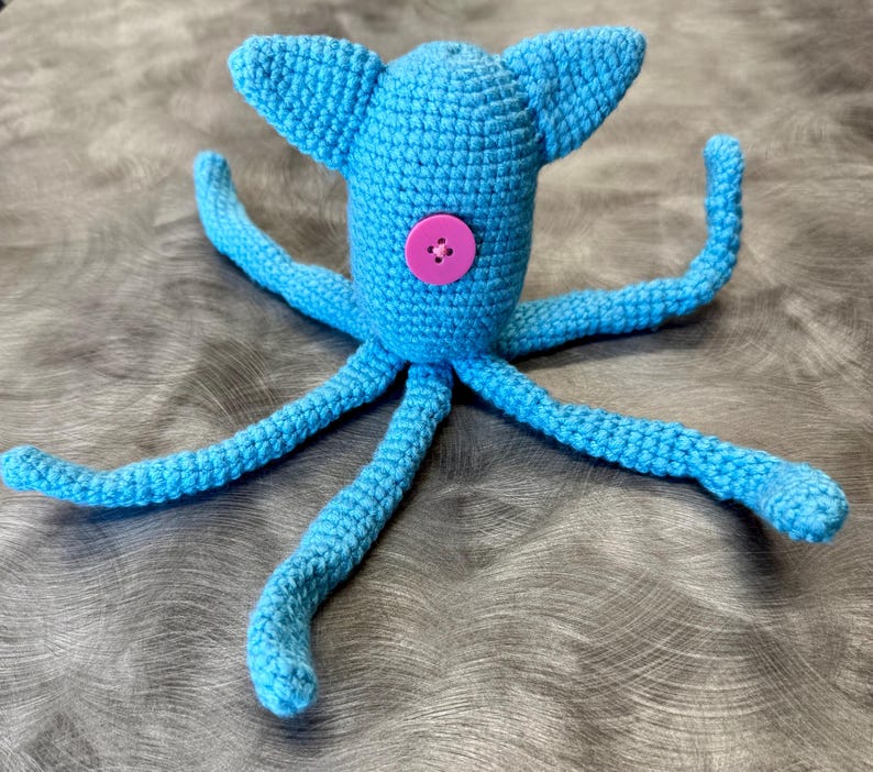 May include: A bright blue crocheted octopus toy featuring a central pink four-hole button on its main body. The creature has two pointed ears at the top and eight long, slender, flexible tentacles extending outward from the base of its head.