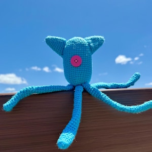 May include: A bright turquoise crocheted creature with a rounded head, two pointed ears, and multiple long, thin tentacles extending outward. A single, circular, hot pink button is securely attached to the center of the creature's head.