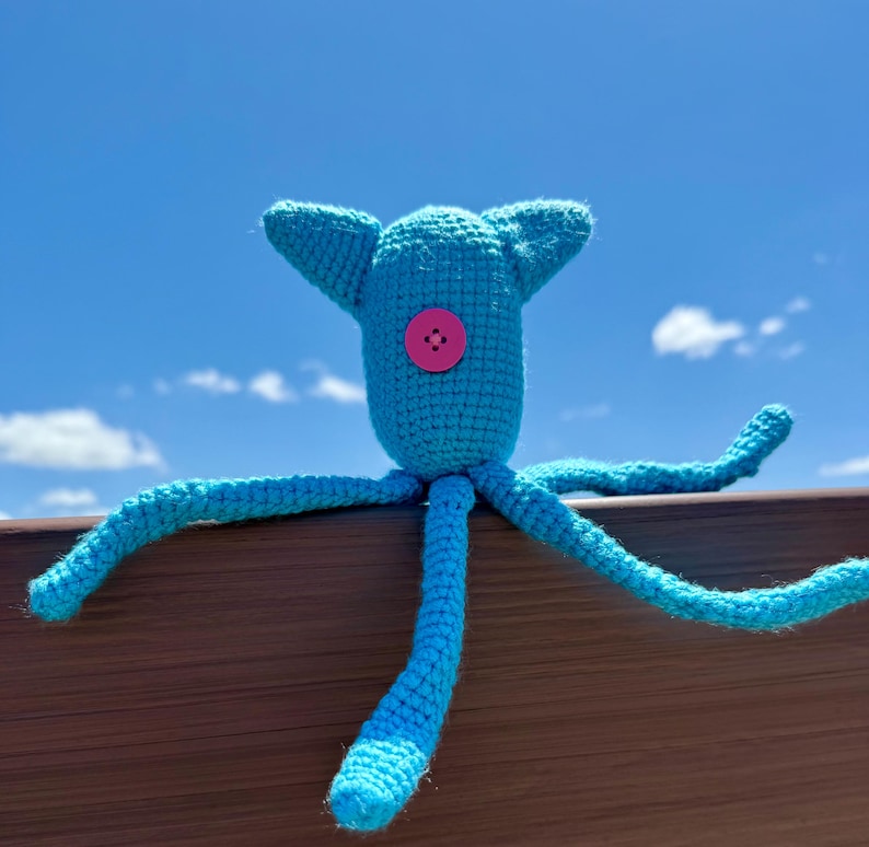 May include: A bright turquoise crocheted creature with a rounded head, two pointed ears, and multiple long, thin tentacles extending outward. A single, circular, hot pink button is securely attached to the center of the creature's head.