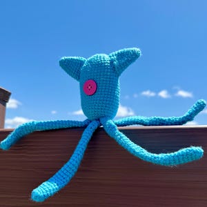 May include: A bright turquoise crocheted creature with a single pink button eye centered on its head. The plush toy features two pointed ears and four long, slender, flexible limbs extending outward from its central body in a star-like shape.