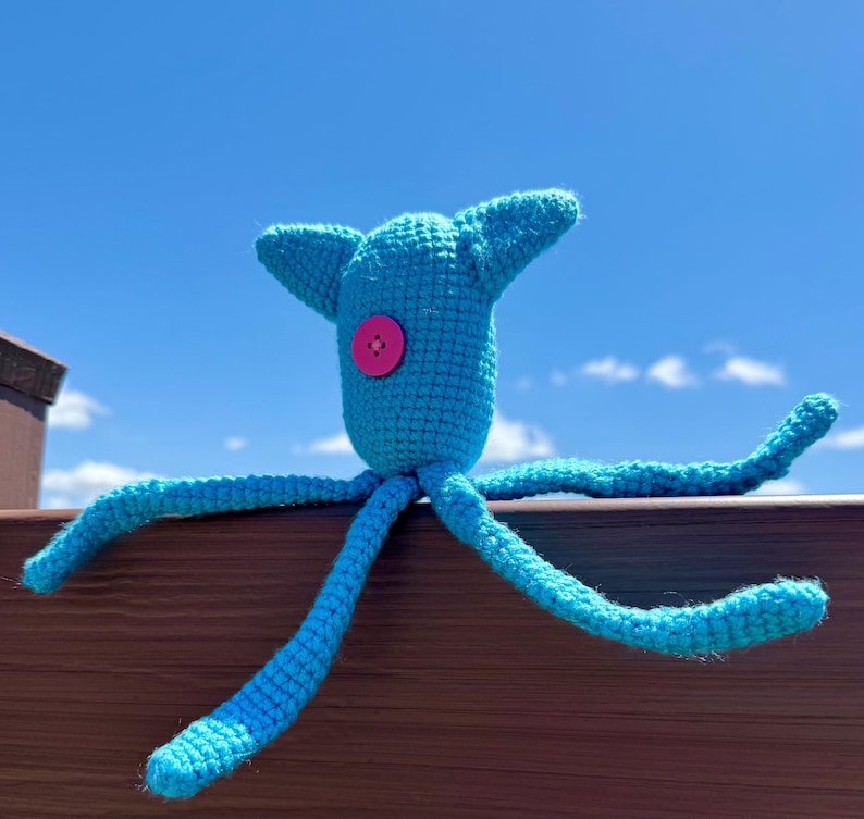 May include: A bright turquoise crocheted creature with a single pink button eye centered on its head. The plush toy features two pointed ears and four long, slender, flexible limbs extending outward from its central body in a star-like shape.