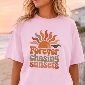 May include: A light pink short-sleeve t-shirt featuring a graphic of a stylized sun and the text "Forever Chasing Sunsets" in shades of orange, yellow, and green. The shirt has a crew neckline and a relaxed, loose-fitting silhouette.