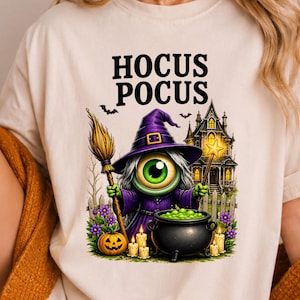 May include: A cream-colored t-shirt featuring a graphic of a cyclops witch in a purple hat holding a broom and wand, with a cauldron, pumpkins, and a haunted house. The text HOCUS POCUS is printed above the graphic in a bold black font.