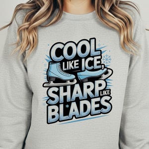 May include: A light gray crewneck sweatshirt featuring a graphic print with the text COOL LIKE ICE, SHARP LIKE BLADES. The design includes two light blue ice skates and snowflake accents, centered on the chest of the long-sleeve pullover garment.