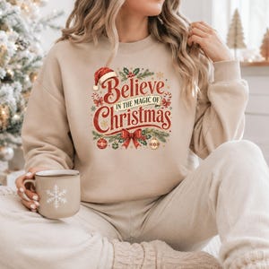 May include: A beige crewneck sweatshirt featuring a graphic print that reads Believe IN THE MAGIC OF Christmas in red and gold lettering, surrounded by illustrations of a Santa hat, candy canes, holly, a red bow, and decorative ornaments.